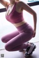 A woman in a pink sports bra top and purple leggings squatting on the floor.